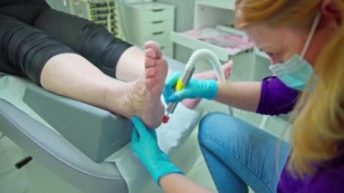 Kozmetik yatakta yatan bir kadın pedikür yaptırır. Güzellik uzmanı topukları yumuşatmak için bir tırnak değirmeni makinesi kullanır. Hastalardan birinin bacakları özel bir bloknotta yatıyor..