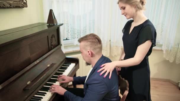 一位音乐家正在为钢琴作曲 他由一名身穿黑色连衣裙的妇女陪同 女人轻轻地抚摸着钢琴家的肩膀和脖子 — 图库视频影像