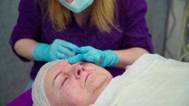 Klinikteki doktor hastaların yüzüne canlandırıcı bir işlem uygular. Küçük delikler kullanarak kadın derisinin altına bir ürün yerleştiriyor. Hastanın yüzü taze kan izleri gösteriyor..
