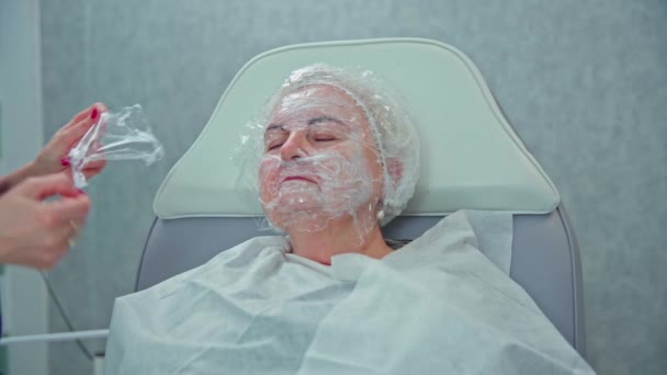 成熟皮肤的全面护理需要去美容院看病 美容师用保湿面膜 病人头上戴着一顶防护帽 她的脸裹在胶卷里 — 图库视频影像