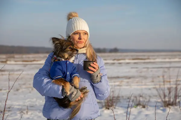 A woman is lost in the wilderness. The young person went for a hike on a winter day and cant find the right way. She looks around with a focused face. She is holding a small dog in her arms.