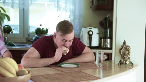 Een Man Zit Aan Keukentafel Een Broodje Eten Achtergrond Ziet — Stockvideo