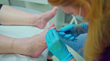 Ön planda bulanıklık. Arka planda kozmetoloji hazırlık ve aletlerinin bulunduğu dolapları ve rafları görebilirsiniz. Bir kozmetikçi müşterilerinin ayak tırnağı üzerinde çalışıyor..