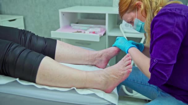 患者は化粧台の上に横たわっている 彼女は足を露出した 美容師は女性の足元で働いています 化粧品学者は足からケラチン化された皮をペンキで取除きます — ストック動画