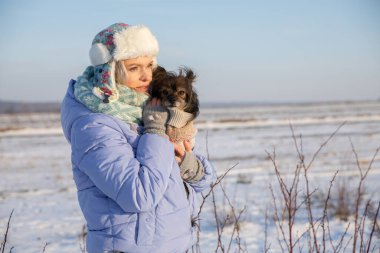 Kış boyunca dışarıda donmuş bir kız. Kollarında küçük bir köpek tutuyor ve ona sarılıyor. Arka planda açık bir alan ve karla kaplı alanlar görebilirsiniz..