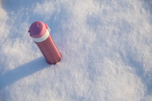 An aerial view of the thermos standing in the snow. The thermos is bright pink in color, with a metallic sheen. The shining sun makes the thermos cast a long shadow on the snow.