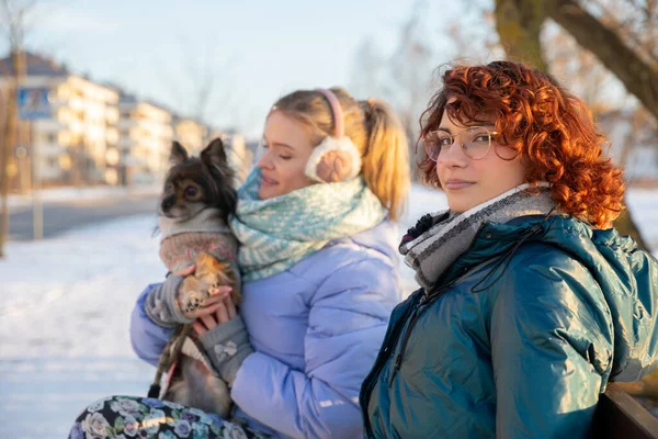 Close-up on the figures of two women sitting on a bench. One of the women is holding a dog on her lap. Both are wearing warm clothes, as it is freezing cold.