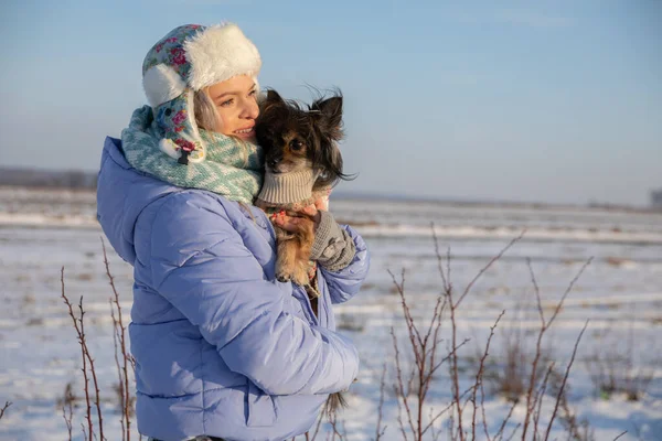 A caring dog owner holds her dog in her arms and cuddles him to keep him from getting cold. It is winter and there is snow on the fields. A distant forest can be seen on the horizon.