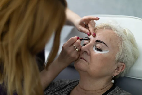 Painting eyebrows at a beautician. A woman enjoys a henna coloring service at a beauty salon. A mature woman sits on a chair in a beauty salon.
