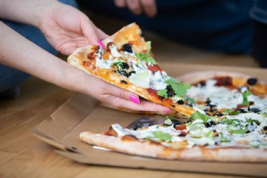 Pizzanın havadan görünüşü. Bazı pizza dilimleri kayıp. Pizzanın yanında oturan biri bir parça alır. Pizzanın üzerinde salam, zeytin ve peynir var..