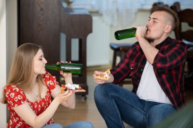 Bir kız ve bir oğlan bir ev partisinde bira içerler. Teslimatla pizza sipariş ediyorlar. Arka planda ahşap bir zemin ve mobilyalar görülebilir..
