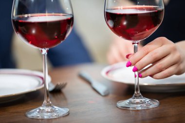 Tahta tezgahta duran iki kadeh kırmızı şarap. Bardaklardan birini bir kadın tutuyor. Kadının tırnakları pembe ojeyle boyanmış..