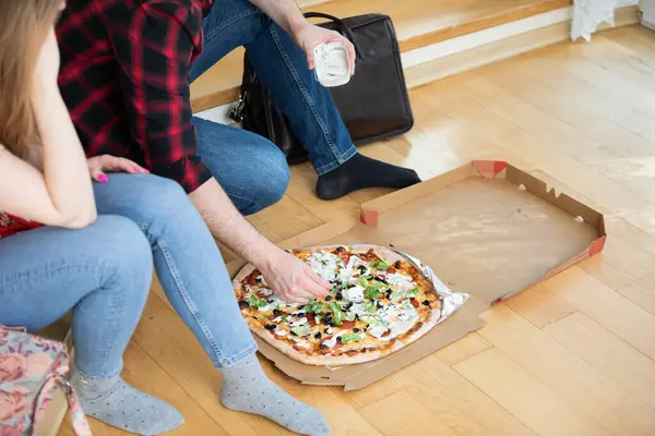 A couple in love has arranged a date. The girl and the boy are sitting on the floor at home and eating pizza. The boy is wearing a plaid shirt.