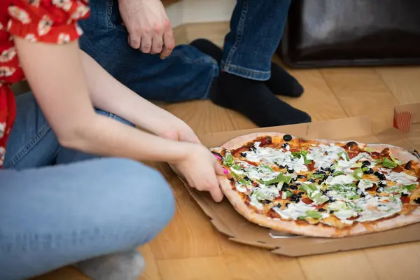 A girl and a boy are sitting on the floor, and a pizza box lies next to them. The girl reaches for a slice of pizza. A couple in love met on a date and are eating pizza.
