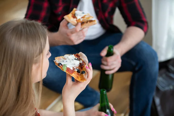View from above. You can see the girls head and hand. She is holding a slice of pizza. In the background you can see the blurred silhouette of the boy, who is wearing a plaid shirt.