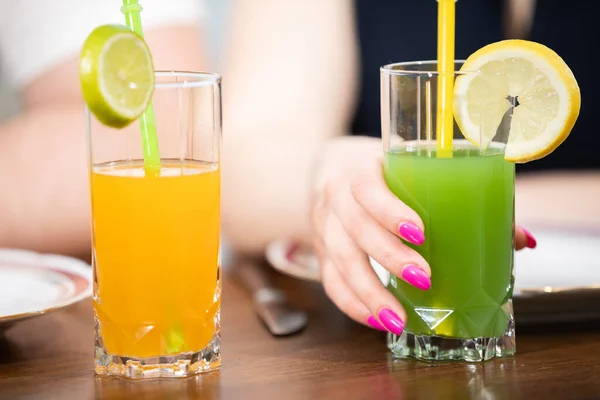 Close-up of two drinks in tall glasses. One of them is orange and the other is green. They both have straws. One of the glasses is held by a womans hand. Woman has pink manicure.