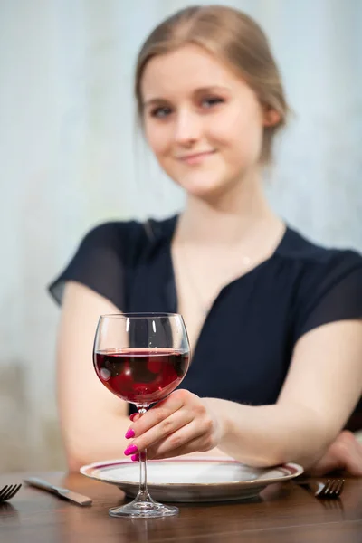 A woman in a black dress sits at a table and holds a glass of red wine. In front of her on the table lies a place setting. The womans figure is slightly blurred.