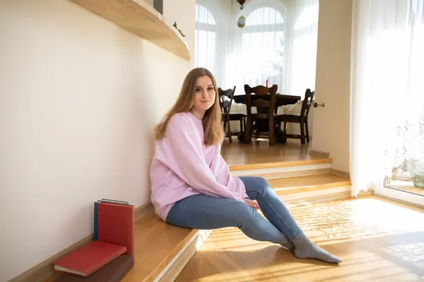 女の子はスポーツスウェットシャツとタイトなジーンズを着ています 彼女は座って部屋の壁に向かっている 背景には 木製のテーブルと椅子があるダイニングルームが見えます — ストック写真