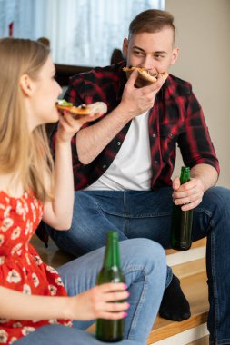 Bir kız ve bir oğlan bir evin zemininde yan yana oturuyorlar. Kız ön planda oturuyor ve vücudu bulanıklaşmış. Pizza yiyorlar ve yeşil camdan bira içiyorlar.
