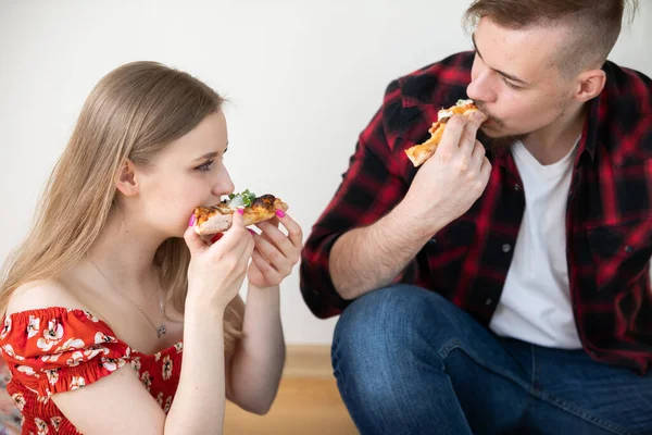 A pair of students ordered a pizza delivery service. They eat it while sitting on the floor. A boy and girl look at each other while eating pizza.
