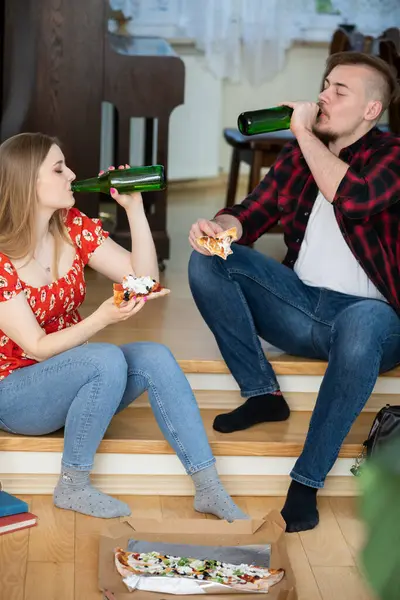 The students held a house party and ordered pizza. A girl and a boy sit on the steps and drink beer. In front of them lies a pizza box.