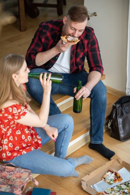 Çift evde akşam yemeği yiyor. Bir adam ve bir kadın merdivenlerde oturmuş pizza yiyorlar. İkisinin de elinde yeşil şişe var. Yerde bir pizza kutusu var..