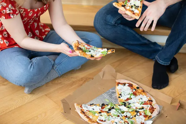 İki kişi bacak bacak bacak üstüne atmış şekilde yerde oturuyor ve yanında bir pizza yatıyor. Her biri bir dilim pizza tutuyor. Arkadaşlar birlikte pizza yiyorlar..