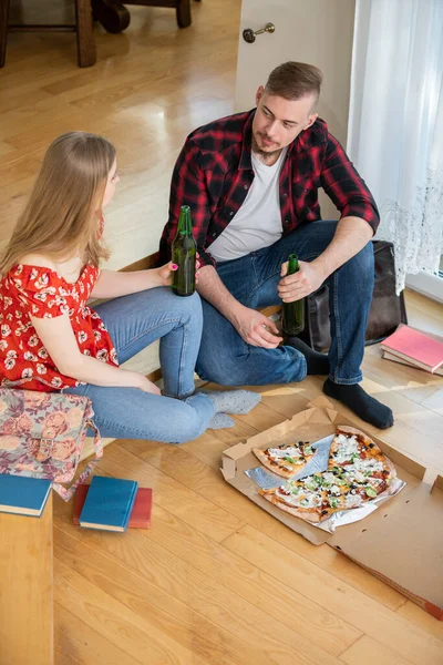 The students met at home to study together. They sit on the floor with books lying around them. The students drink beer and eat pizza while studying.