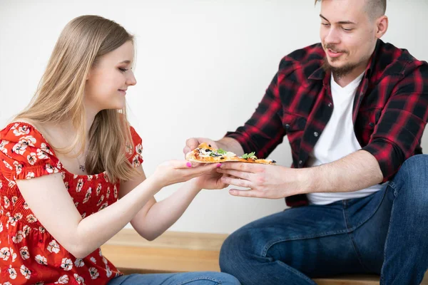 A man and a woman sit on the floor and eat pizza. They are sitting close to each other. The man hands a piece of pizza to the woman, and she takes it.