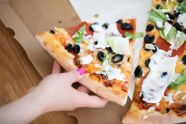 A close-up of a pizza in a box, lying on the floor. A womans hand reaches for a piece of pizza. Pizza with black olives, tomato and garlic sauce.