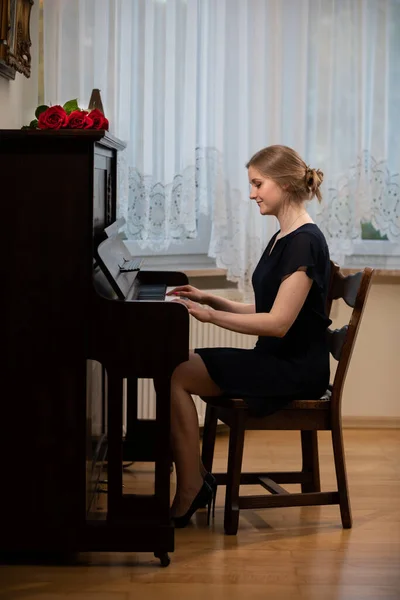 An elegant woman plays the piano in the living room. She is wearing a black dress and high heels. A bouquet of red roses lies on the piano.