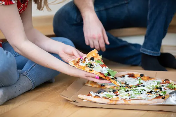 A girl and a boy have ordered pizza for dinner. The couple is sitting on the floor with a pizza in a box in front of them.