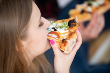 Bir kadın bir dilim pizza tutuyor. Isırıyor. Büyük bir yakın çekim. Kadının yüzü profilden görünüyor. Bir sürü malzemeli renkli pizza..