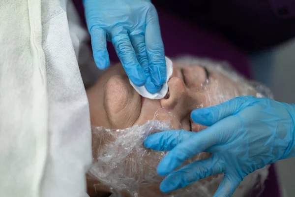 Close-up of the face of a mature woman. The face is partially covered with a film. This is part of a beauty treatment. A beautician wearing disposable gloves cleanses the skin of the face with a gauze
