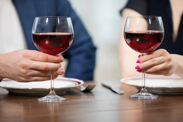 Two glasses of red wine stand on the table. A man and a woman are holding their glasses. In the background you can see blurred figures of a man and a woman sitting at a table.
