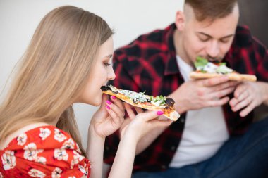 Ön planda genç bir kadın profili var. Saçları açık. Kadın bir dilim pizzayı ısırıyor. Arka planda bir adam görülebilir, vücudu bulanıktır. O da yiyor.