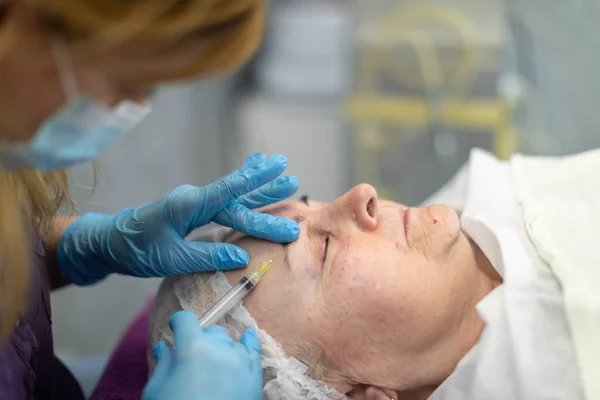 Side view of the face of a lying woman. She has her eyes closed. A nurse gives the woman subcutaneous injections near her eyebrows.