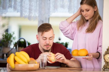 Bir adam mutfak masasında oturmuş sandviç yiyor. Arka planda perdeli bir pencere, mutfak tezgahı, lavabo ve çeşitli mutfak aletleri görebilirsiniz. Bir kase meyve standı yanında.