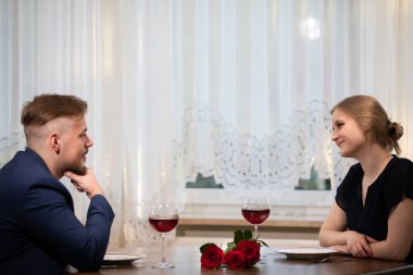 Bir kadın ve bir erkek yüz yüze bir masada oturuyorlar. İkisi de birbirine bakıyor. Masada bir buket kırmızı gül ve iki bardak kırmızı şarap var. Arka planda bir...