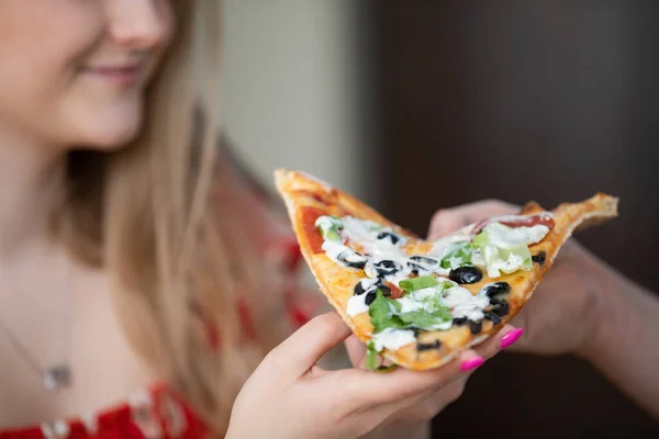 Pizza with rucola, black olives and salami slices. There is a white garlic sauce on the pizza slice. The figure of a young woman can be seen in the background.