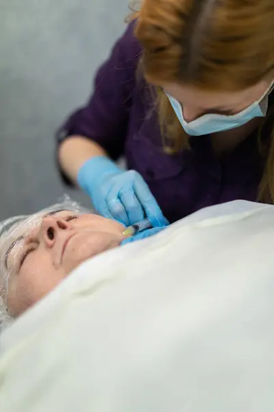 A patient of an aesthetic clinic is lying on a bed. She is covered with a white coverlet. Next to her sits a nurse who performs a needle puncture with a syringe. She injects the skin of her face.