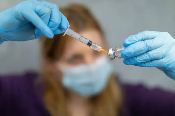 Blurred foreground. The sharpened foreground shows hands wearing disposable gloves. One hand is holding a vial of medicine, while the other is acquiring the drug using a syringe with a needle.