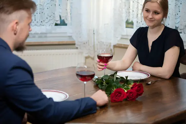 A girl in a black dress sits at the table. There is a bouquet of red roses on the table. A boy in a blue jacket sits opposite the girl.