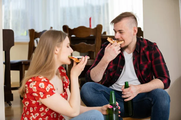 Hungry young people at a party are eating an ordered pizza. A girl and a boy sit across from each other. They both take bites of pizza slices. In the background you can see blurred chairs.