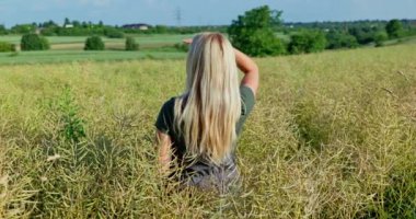 Profesyonel bir kadın çiftçi, gri tulumlar ve kısa kollu bir zeytin gömleği giyer. Sırtı kameraya dönük durur. Olgunlaşan kolza ve buğday tarlalarına bakar..