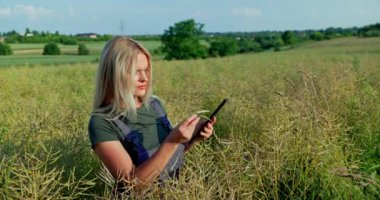 Gevşek, uzun sarı saçlı bir kadın çiftçi olgunlaşan kolza tohumu tarlasını inceliyor. Görünüşünü kalite kontrol için bir tablet uygulaması kullanarak online resimlerle karşılaştırıyor..