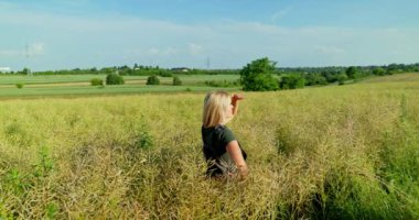 Zeytin rengi tulumlar ve kısa kollu bir gömlek giyen profesyonel bir kadın çiftçi, olgunlaşan tecavüz tohumu tarlalarının arasında titizlikle mahsullerini tarıyor..