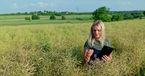 輝く真昼の日差しの中で ブロンドの髪を流すプロの女性農家は 今後の収穫を予想して満たされた熟したレイプ作物を検査します この年で喜ぶ — ストック動画