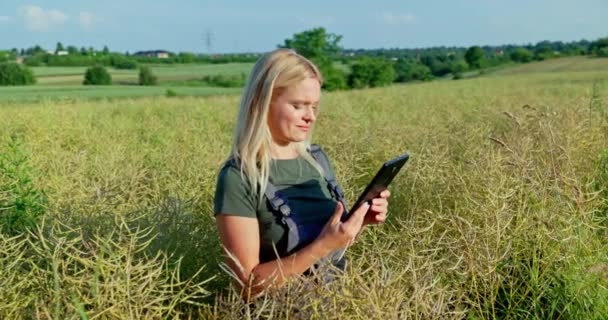 明るい正午の太陽に ブロンドの髪を持つ喜ばしい農夫は 繁栄した収穫を予測して 彼女の熟したレイプ畑を検査します — ストック動画