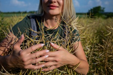 The woman is engaged in agriculture. She owns a rapeseed field and is checking this years harvest. She is an experienced farmer, caring for her crops in a vast field. She has blonde hair and smiles clipart